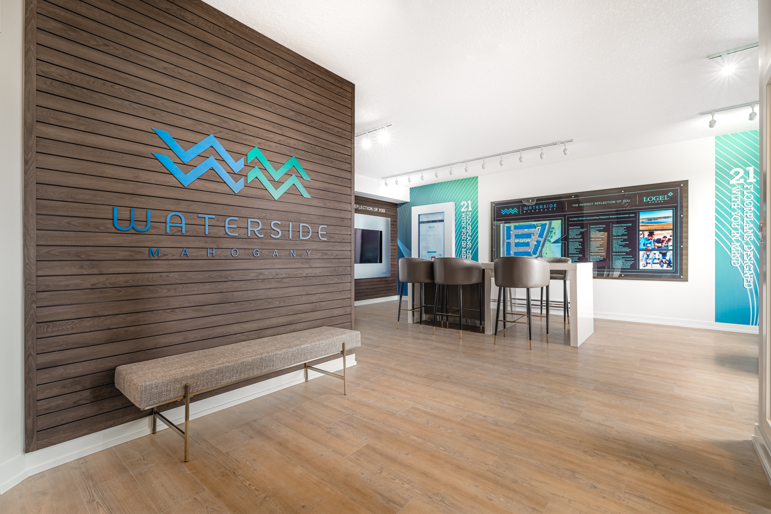 Waterside at Mahogany Sales Centre interior featuring a wooden feature wall with the logo, display island, and large community map with two interactive screens on either side.